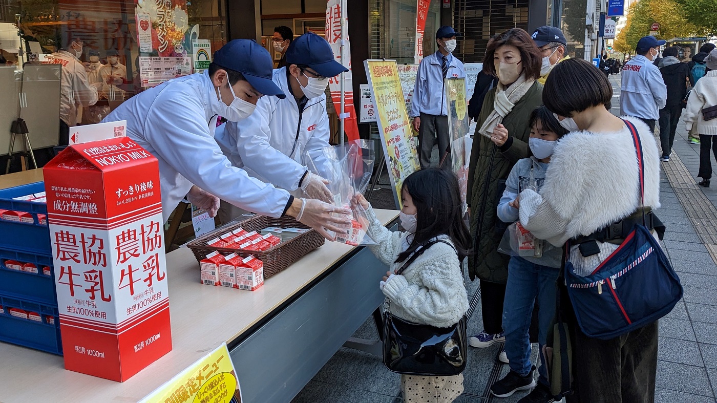 JA東京アグリパークの店先で家族連れに牛乳を手渡す全農職員